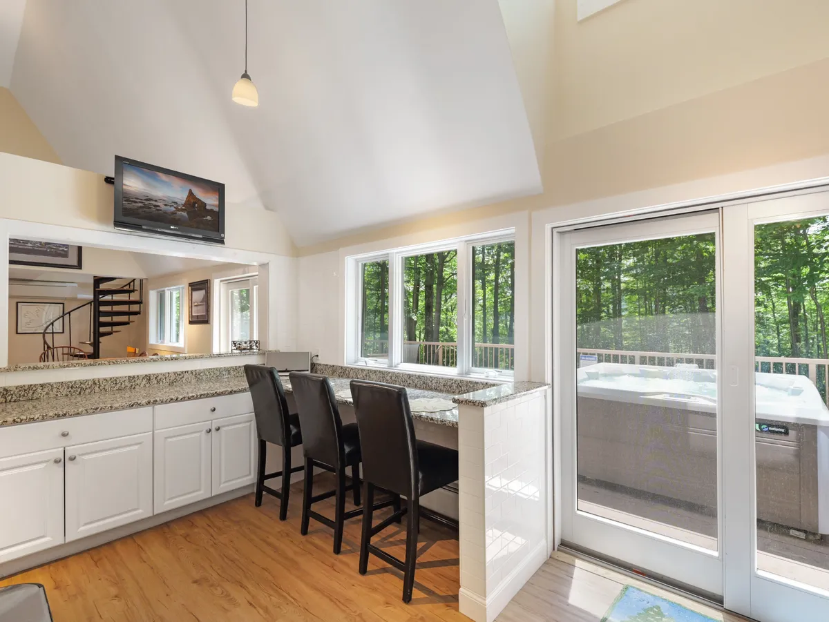 A view from inside the home looking through large windows at the outdoor hot tub. The image includes a cozy granite countertop with black bar stools, hinting at the home’s inviting social spaces and the allure of après-ski relaxation.