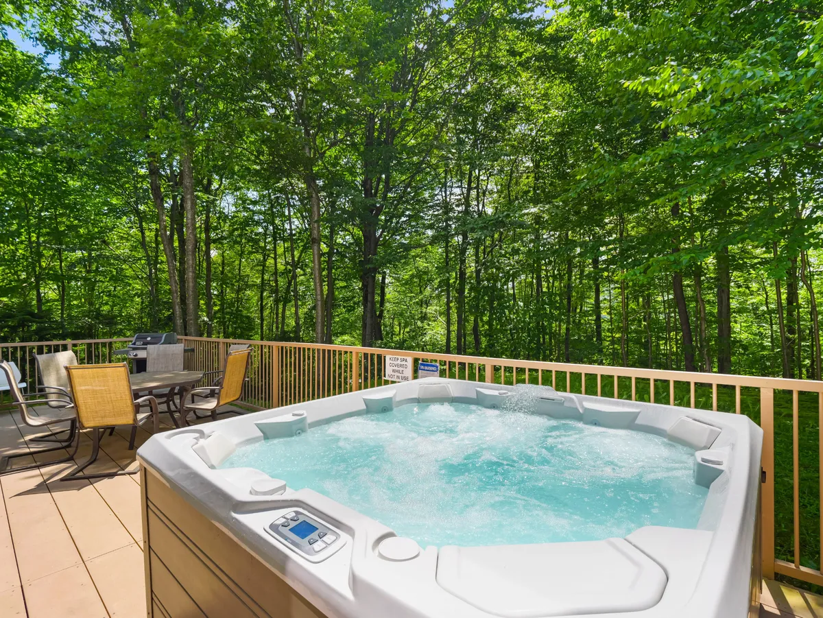 An inviting exterior view featuring the bubbling hot tub, outdoor seating area, and lush green trees surrounding the property. The tranquil setting encourages relaxation and socializing outside, with room for both adults and children to enjoy the fresh mountain air.
