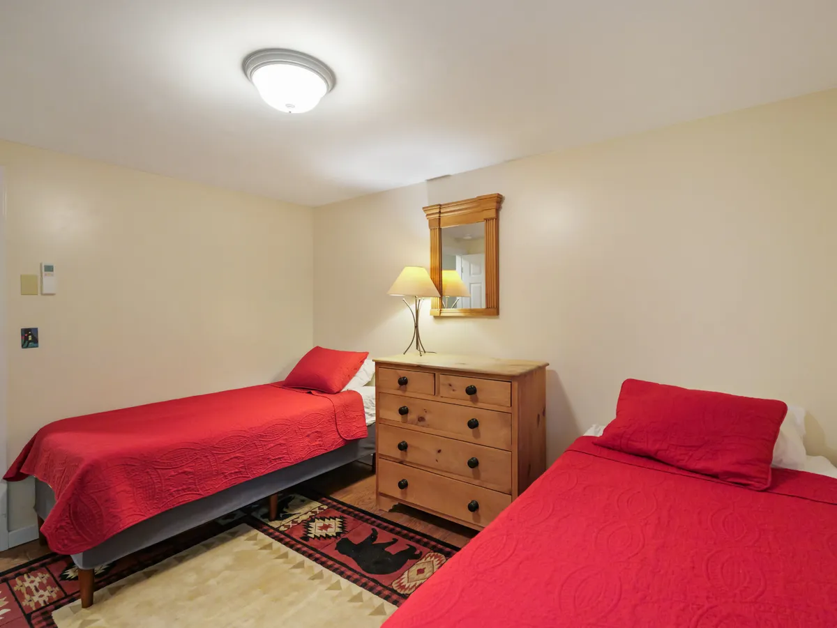 Two twin beds with matching red bedspreads are arranged in a warmly lit room, complemented by a wooden dresser, lamp, and wall mirror. The décor features natural wood elements, and the space feels both inviting and functional for guests.