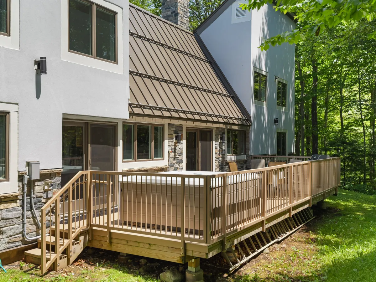 This exterior image showcases the expansive deck of the 76 House surrounded by lush greenery and the home's welcoming architecture. You can see large windows that bring in natural light, the inviting design of the roof, and the tranquil environment provided by the neighboring forested landscape.