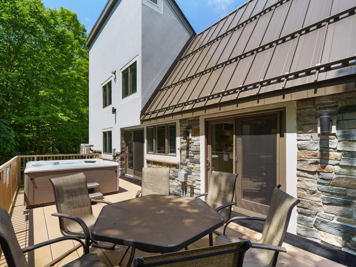This exterior image showcases a hot tub set on a deck, complete with an outdoor dining table and a partial stone wall. The area is surrounded by nature, creating a serene environment for relaxation and socializing after a day on the slopes.