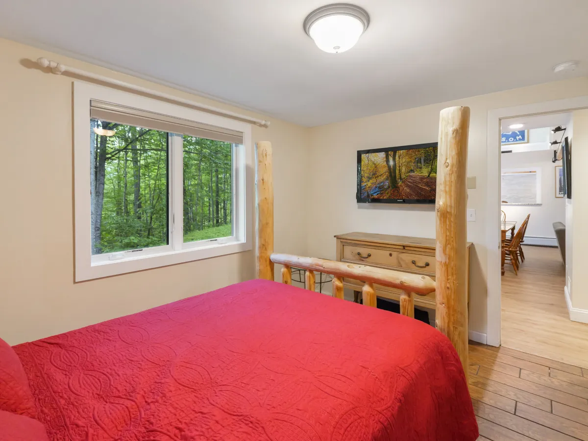 A bedroom with a bold red bedspread, large window allowing in natural light, wooden dresser, and tasteful artwork on the wall. The space exudes a sense of tranquility, with gentle lighting and thoughtful decor choices creating an atmosphere ideal for relaxation.