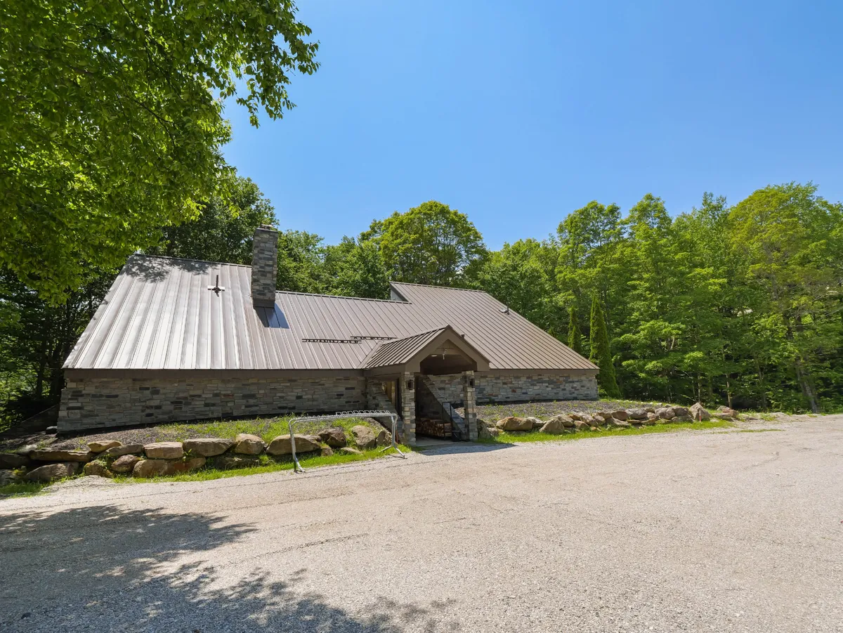 An exterior view featuring a stone facade, metal roof, and wooden entrance, surrounded by a serene natural environment. The image evokes a sense of arrival and warmth, inviting guests to step inside and make themselves at home.