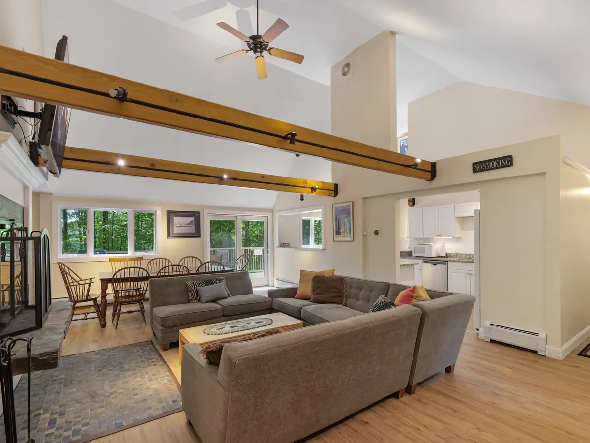 The image showcases a spacious, sunlit living and dining area with inviting sofas, a large rustic dining table, exposed wooden beams, and expansive windows that frame the natural surroundings. The warm, welcoming atmosphere encourages guests to gather, relax, and enjoy each other's company after a day on the slopes.