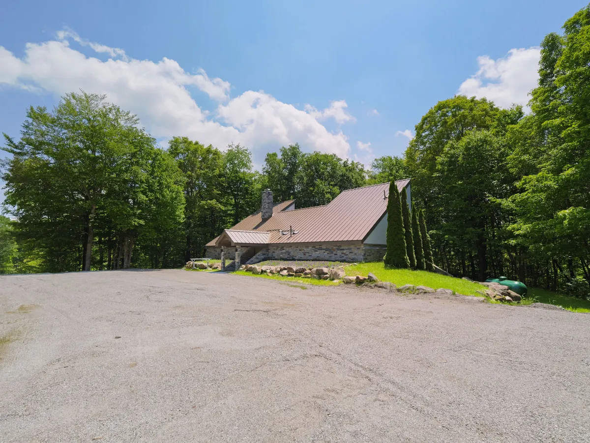 A serene exterior shot featuring the stone-clad facade of the house with its metal roof and surrounded by tall trees. The gravel driveway and natural setting evoke a sense of seclusion and provide an immediate connection to the outdoors and nearby mountain trails.