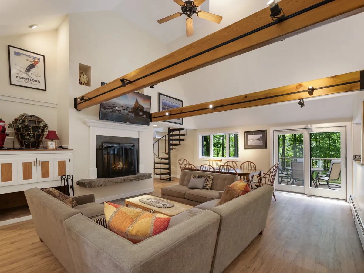 A spacious open-concept living and dining area featuring a cozy wood-burning fireplace, plush sofas, a large dining table, and a prominent spiral staircase. Panoramic windows fill the space with natural light and likely offer views of the mountains.