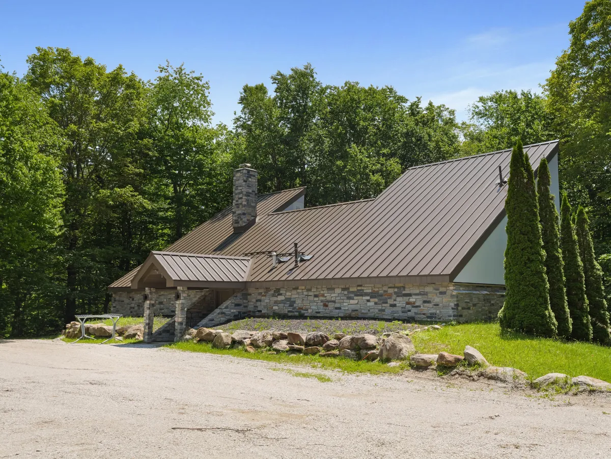 A wide exterior shot of the house set against a backdrop of tall coniferous trees, showcasing the modern architecture, natural stone facade, and metal roof. The tranquil setting conveys the privacy and immersion in nature that the 76 House offers.