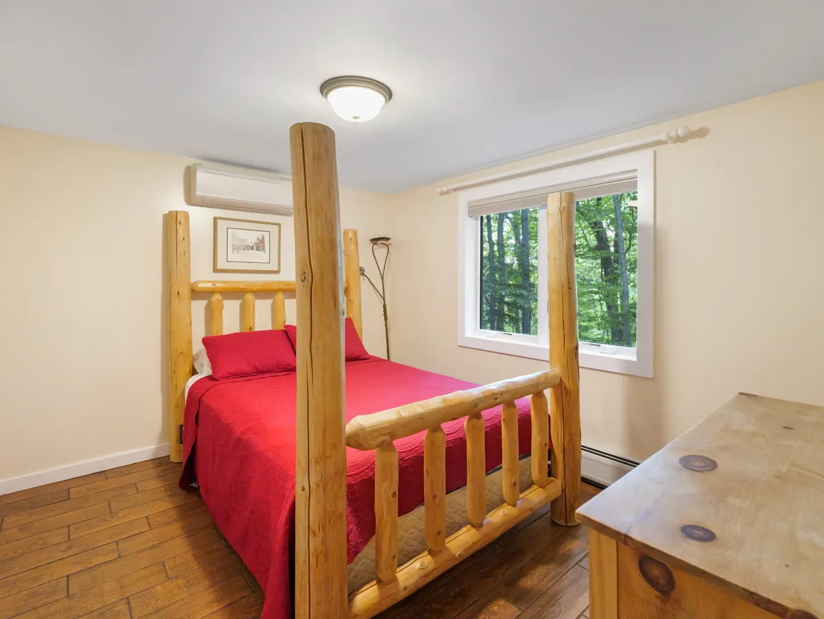 A cozy log bed with inviting red bedding sits next to a window that offers a view of the outdoors. The space feels warm and comfortable, with natural wood accents that hint at its mountain setting. Soft lighting and rustic decor give the bedroom a restful and serene atmosphere.