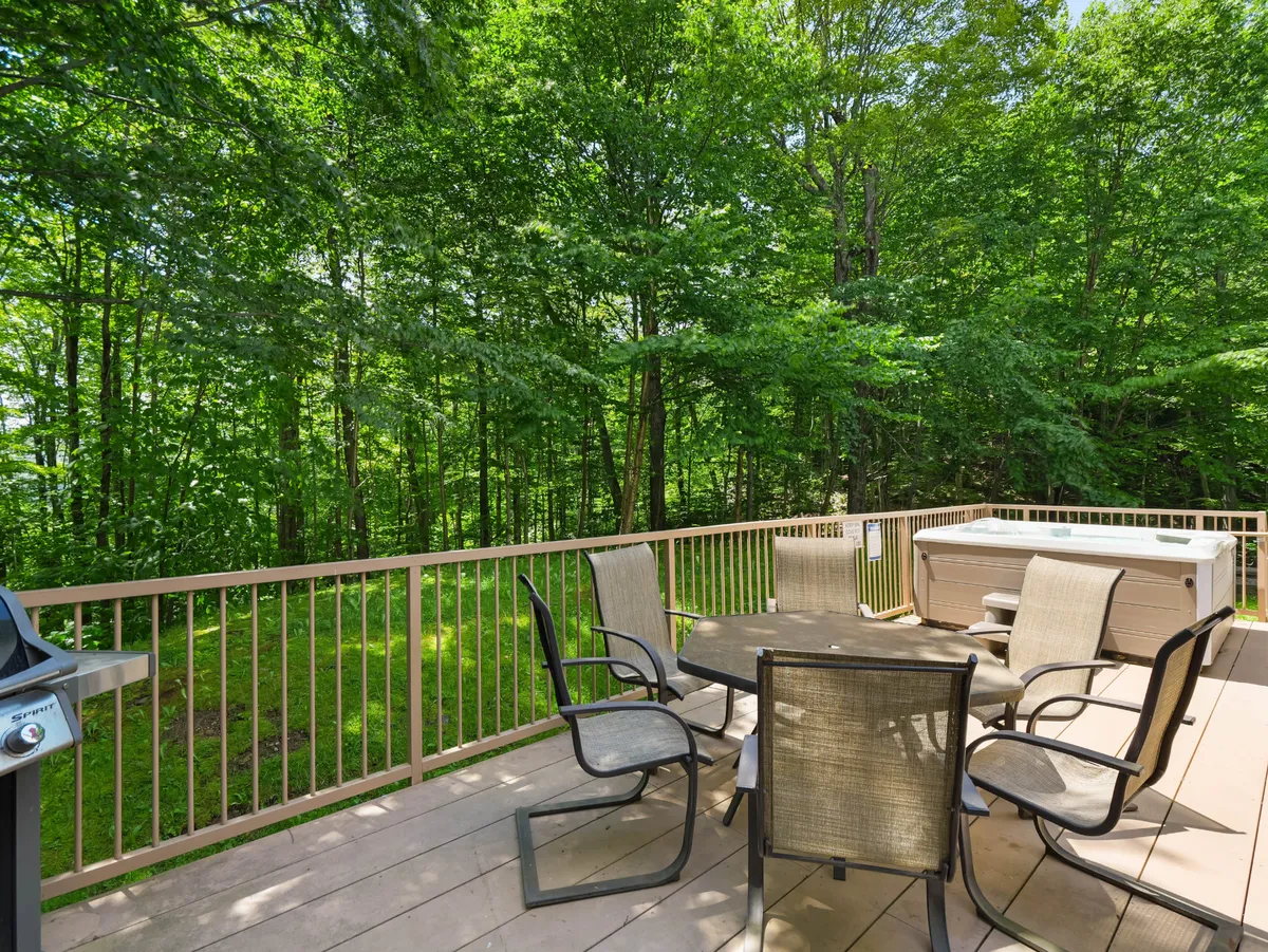 An inviting exterior view featuring an outdoor dining table, hot tub, barbecue grill, and a backdrop of lush trees. The setup suggests relaxation and social activities, with the hot tub and grill highlighting the home's amenities for summer evenings and post-adventure unwinding.