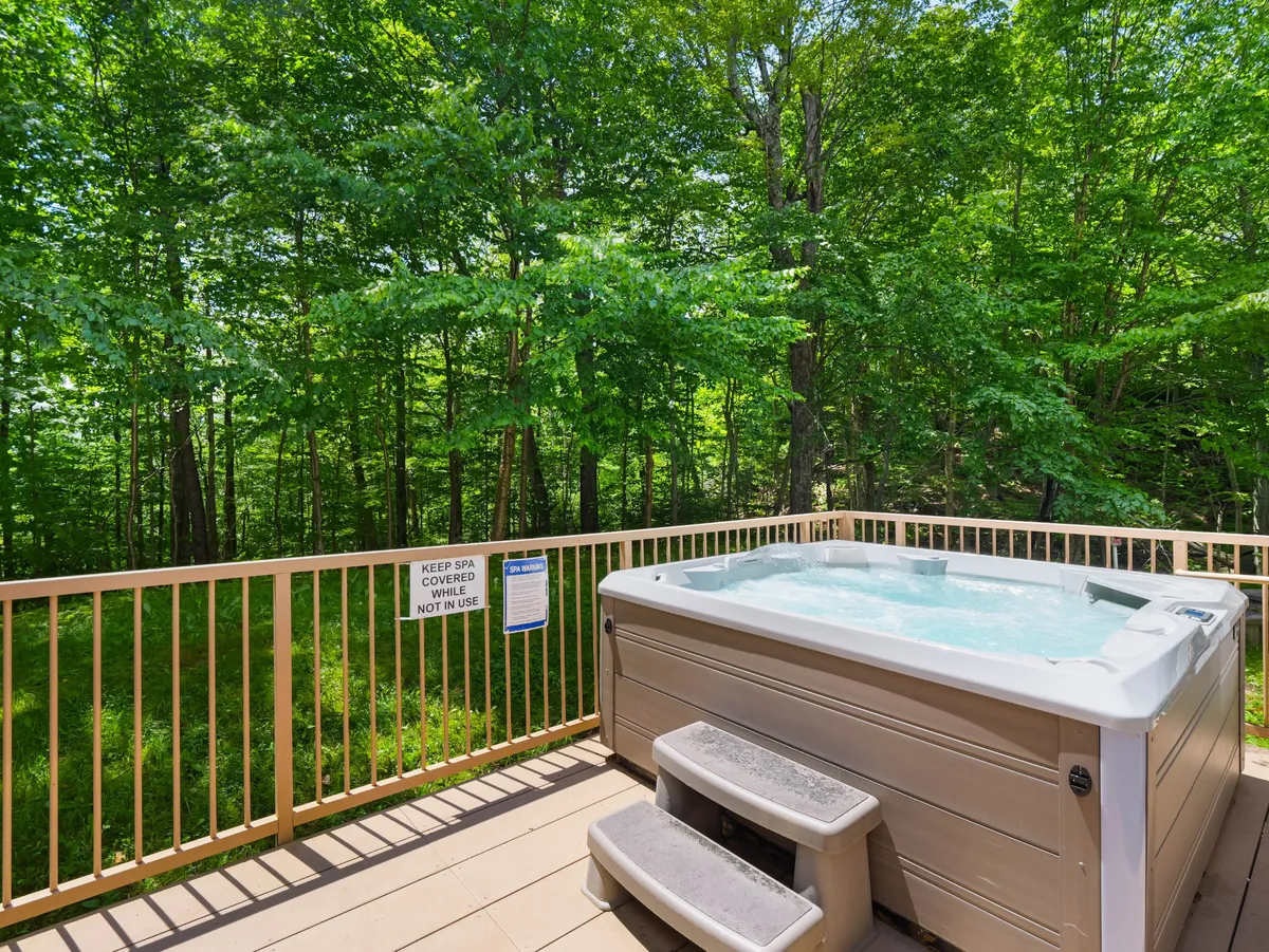 An exterior shot of a hot tub set on a wooden deck surrounded by trees, providing a secluded, tranquil setting. The hot tub is featured as the primary focal point, inviting guests to relax while taking in the natural beauty of the mountain environment.
